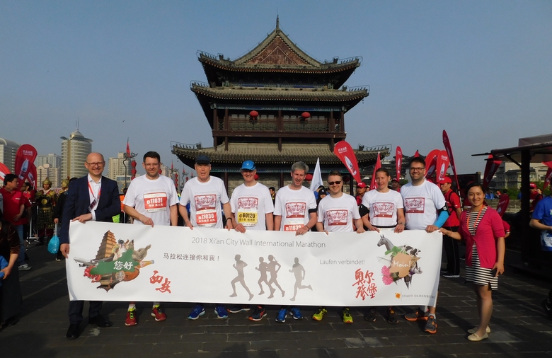 Sieben Läuferinnen und Läufer halten ein Transparent. Rechts und links stehen ein Mann und eine Frau, im Hintergrund ein tempelartiges Gebäude.
